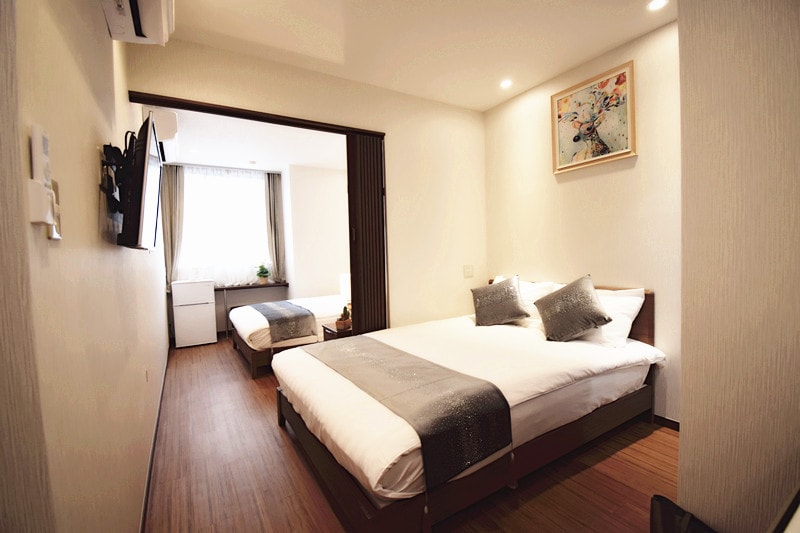 A well-lit room with two beds, one large bed positioned in the foreground and a smaller bed visible in the background. A soft blanket and decorative pillows are arranged on the large bed. A window allows natural light to fill the space, complemented by minimalistic decor.