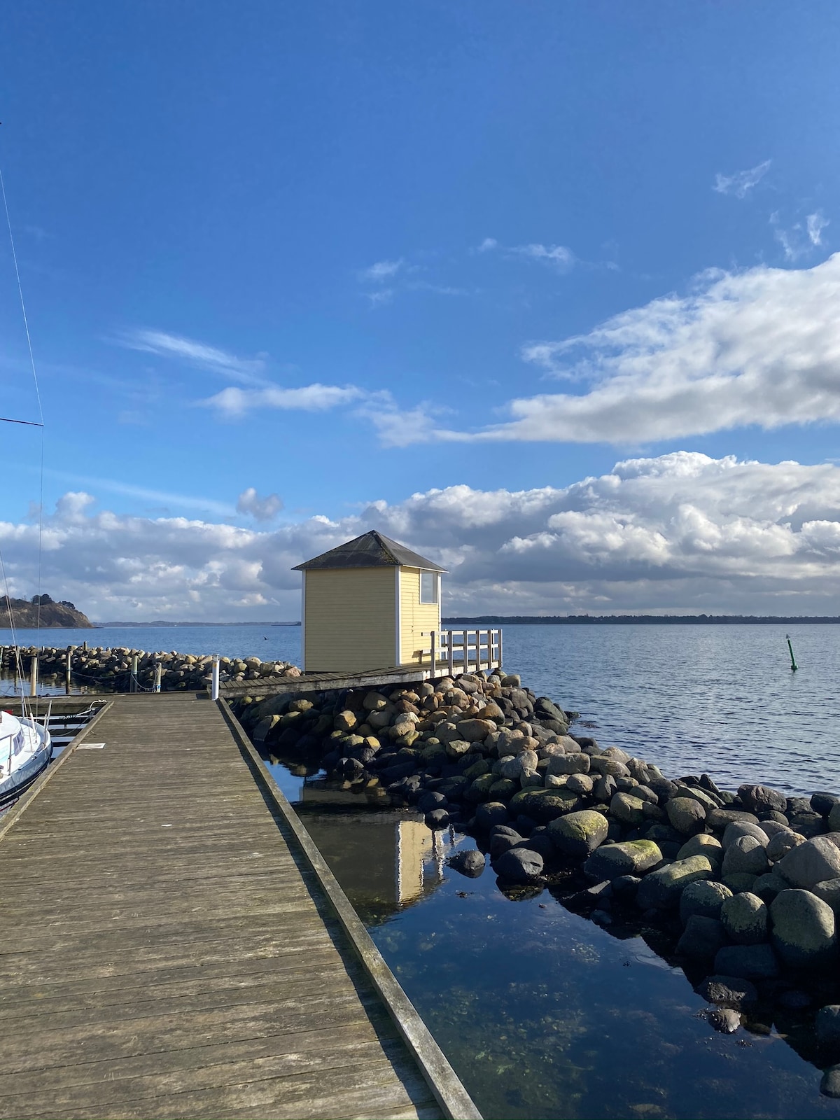 A small yellow building is positioned at the end of a wooden dock. Surrounding it are smooth stones along the water's edge, reflecting the blue sky and fluffy white clouds. The serene body of water stretches out, creating a peaceful coastal scene.