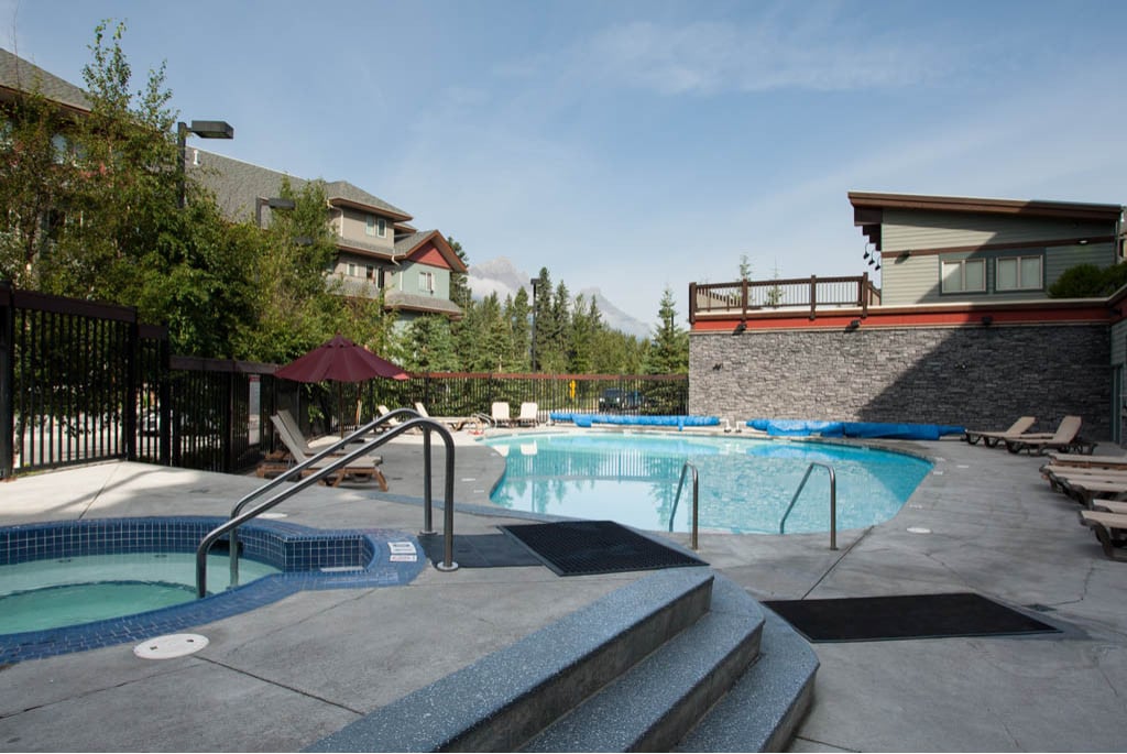 An outdoor pool area is surrounded by trees and buildings, with a large swimming pool and three hot tubs positioned nearby. Lounge chairs line the pool's edge, offering a place for relaxation under the open sky and scenic mountain views.