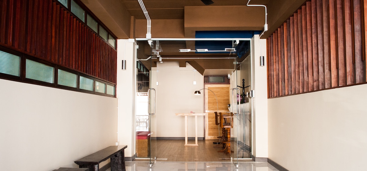 An open layout is presented with a glass sliding door, leading into a room with a wooden desk and chairs. Natural light filters in through large windows, highlighting the minimalistic style of the space with smooth flooring and wooden accents on the walls.