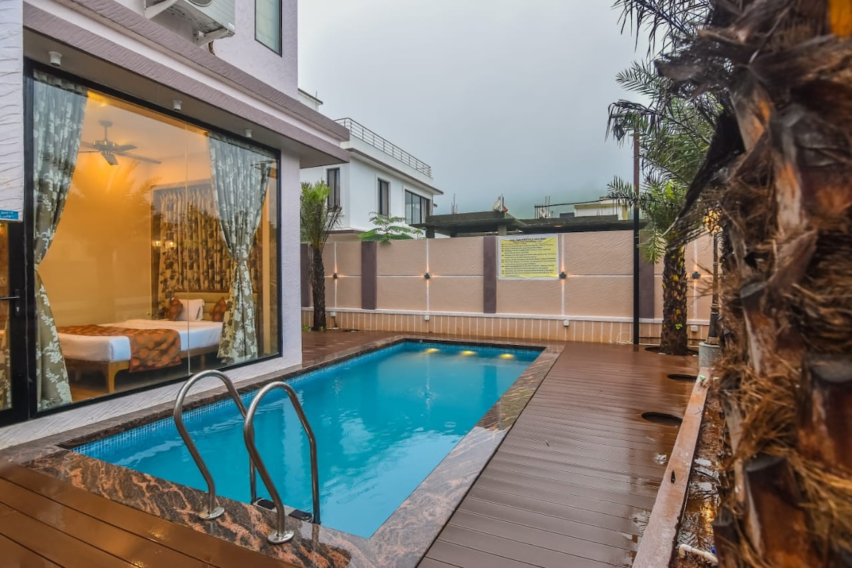 A private pool is surrounded by a wooden deck, offering a serene outdoor space. Large glass doors provide views into a well-appointed bedroom with light-colored linens and patterned curtains. Lush greenery accents the area, enhancing a natural feel in the tranquil setting.