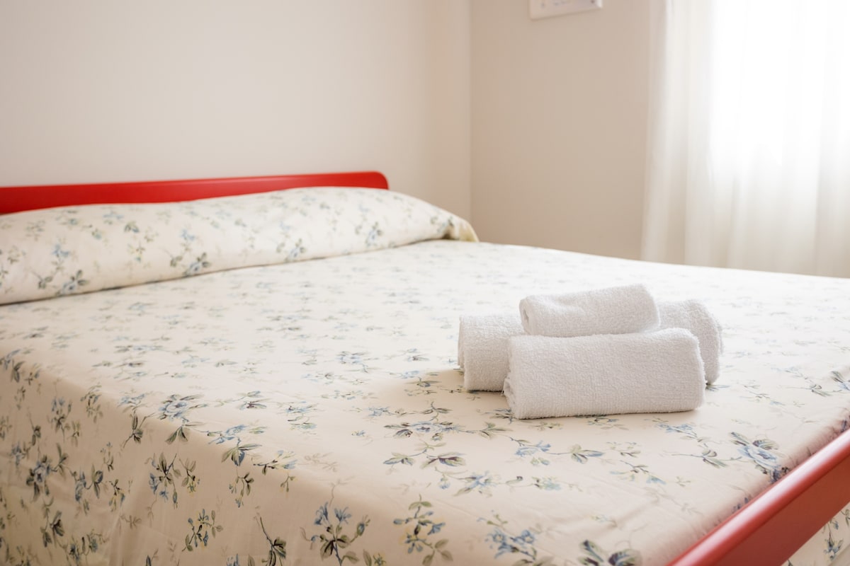 A spacious bed is dressed in a floral-patterned duvet cover, complemented by neatly rolled white towels placed on its surface. Natural light enters through a nearby window, enhancing the soft, neutral color palette of the room.