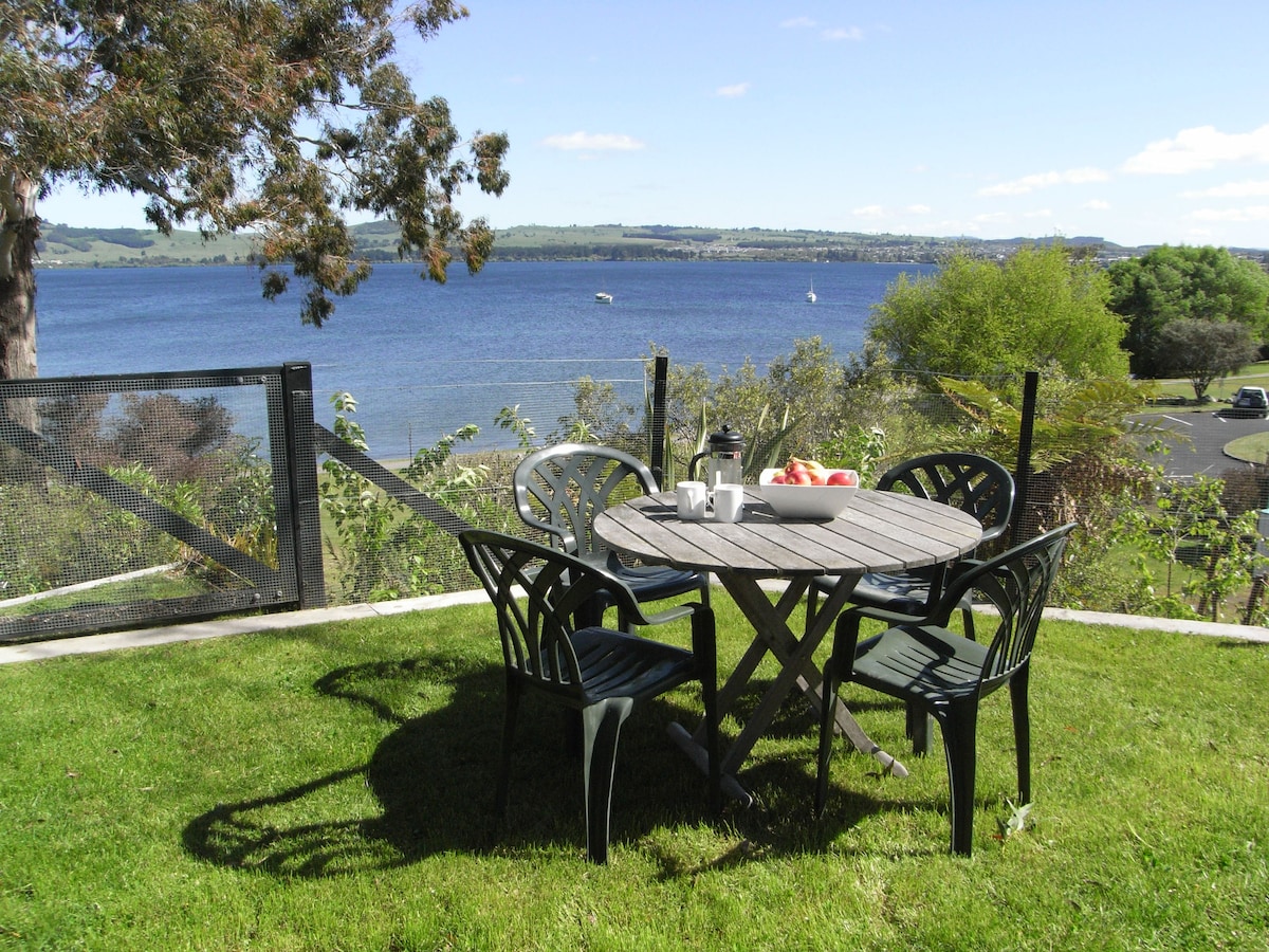 A round wooden table set with cups and fresh fruit is surrounded by four black chairs on a grassy area. A view of the lake is visible in the background, framed by trees and a clear blue sky.