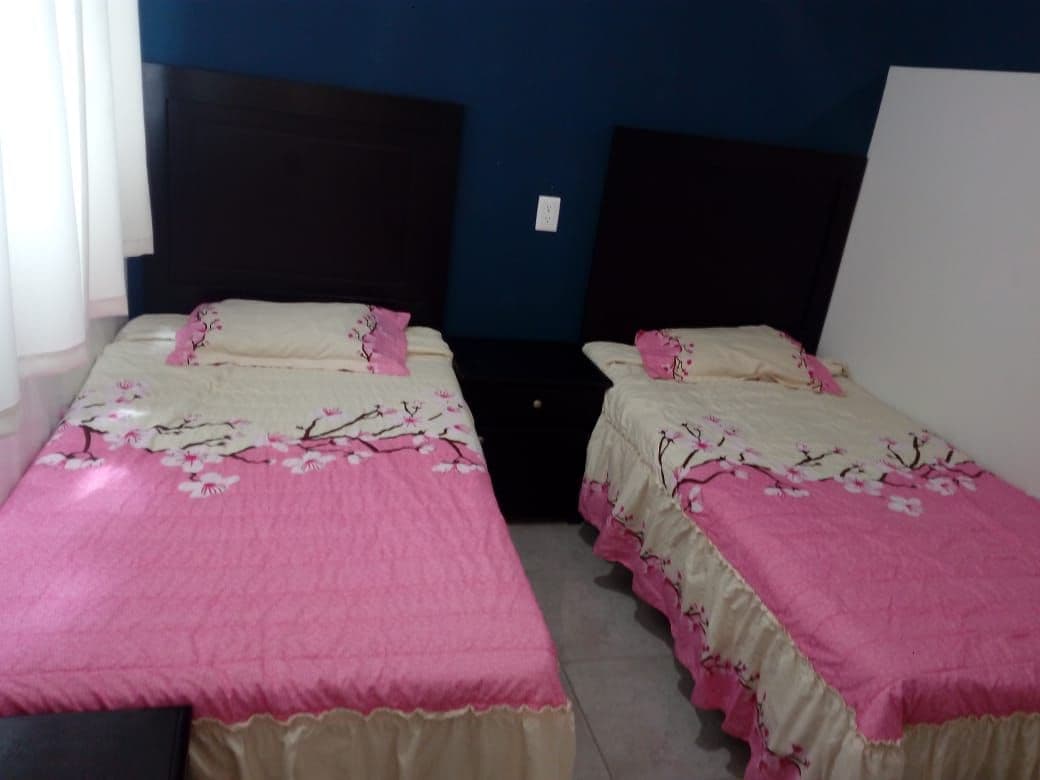 A sleeping area is furnished with two beds, each with floral-patterned bedding in shades of pink and cream. A nightstand is positioned between the beds. Natural light is allowed in through a window with light-colored curtains.