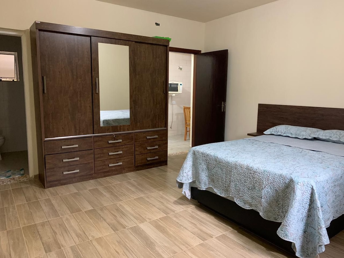 A spacious sleeping area features a large bed covered with a light blue quilt. A wooden wardrobe with multiple drawers and mirrors is positioned against the wall, reflecting soft lighting. The floor is tiled, providing a clean and modern appearance.