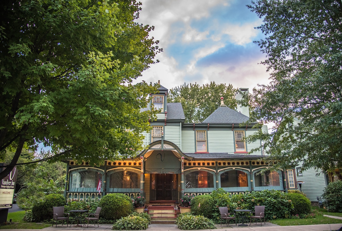 The exterior of the Vandiver Inn features a charming, multi-story structure painted in light green. Lush greenery surrounds the entrance, with a welcoming porch adorned with seating. The building's architectural details are highlighted by large windows and decorative trim, complemented by a serene sky.