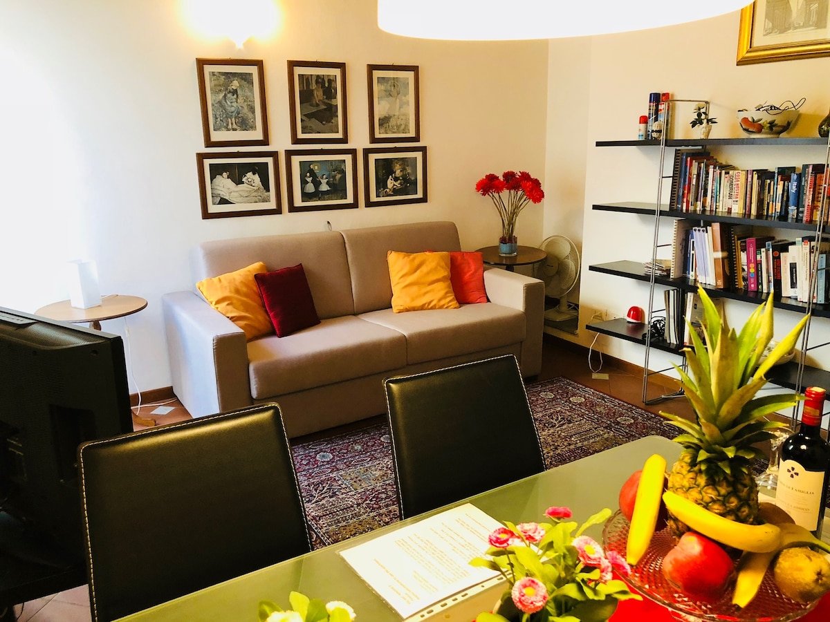 A living area is displayed, featuring a neutral sofa adorned with colorful pillows. Wall art consisting of framed prints adds character. A bookshelf filled with various books is present, complemented by a dining table set with fruit and decorative flowers.