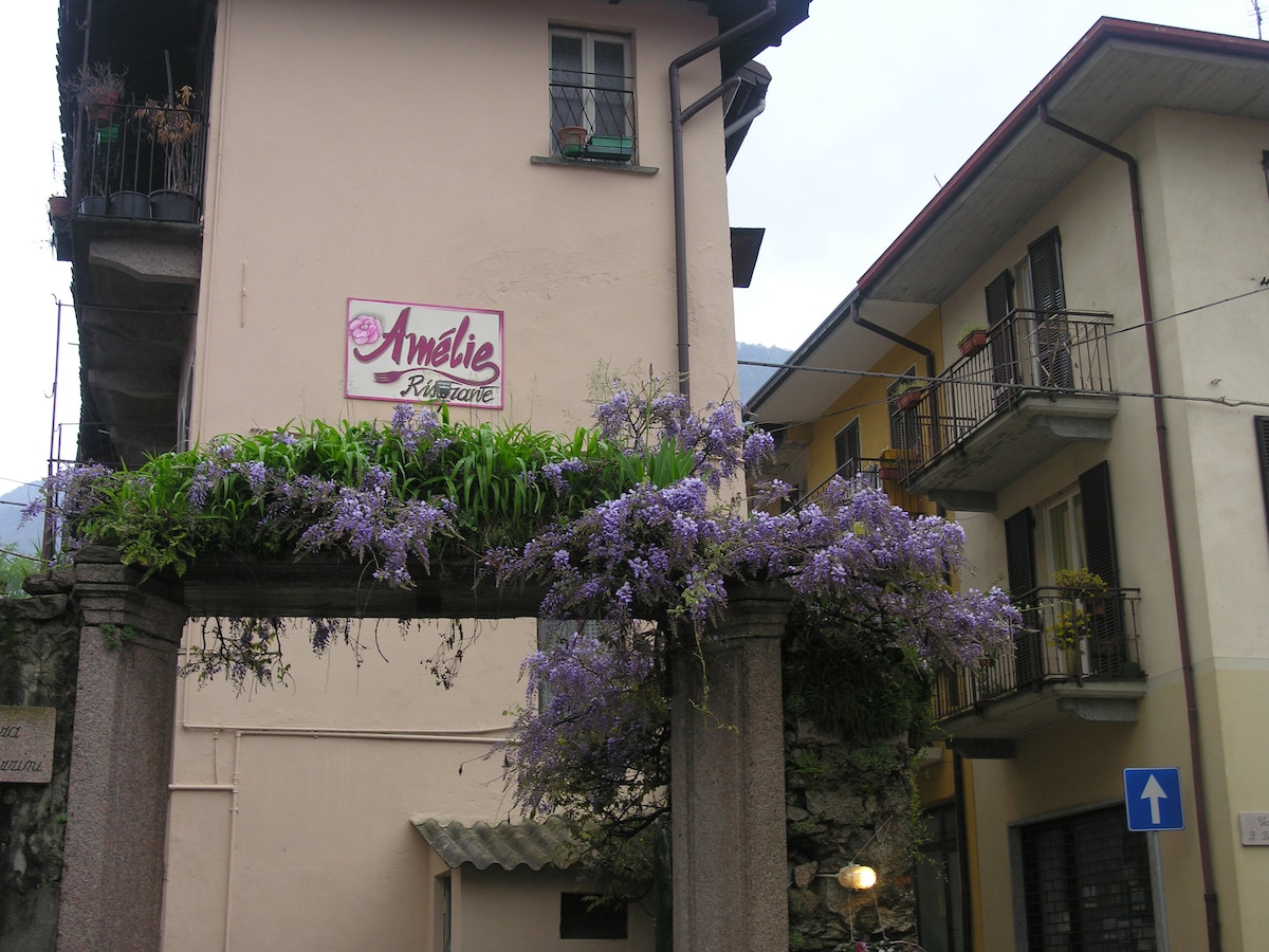 The exterior of L'Affittacamere Ristorante Amèlie is showcased, with a prominent sign featuring the name 'Amèlie Ristorante'. A climbing wisteria blooms above a stone archway, leading to the entrance. Two balconies are visible on the upper floors, adorned with potted plants.