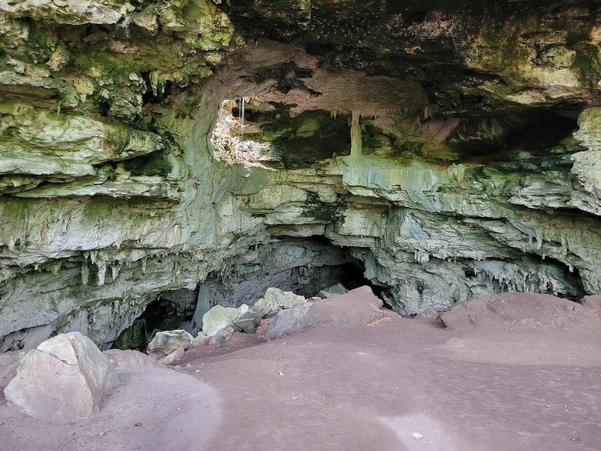 A natural cave entrance is visible, with weathered rock formations showcasing a range of green hues. Sandy terrain leads into the darker interior of the cave, where additional rock layers are discernible. The sunlight filters through the cave opening, highlighting the unique textures of the stone.