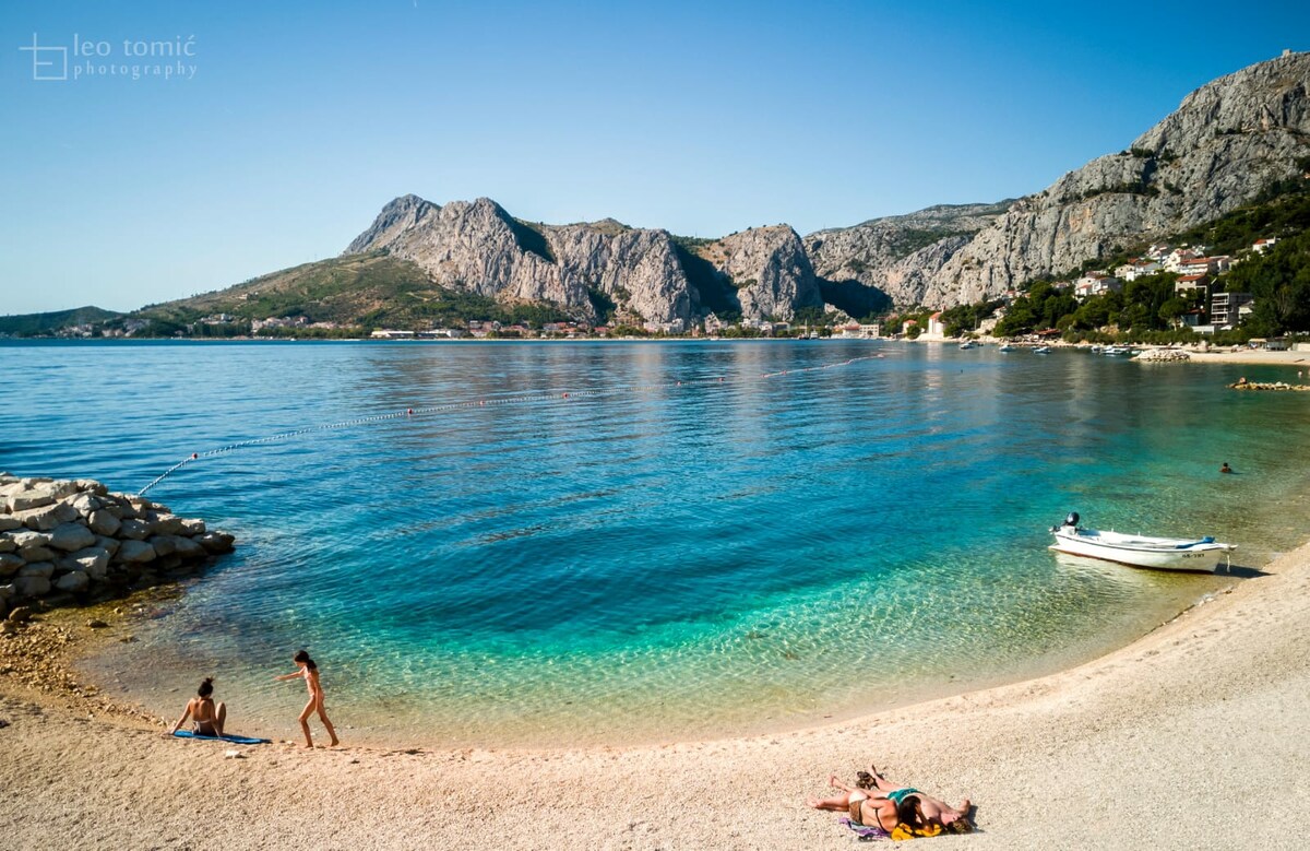 A serene beach scene shows calm turquoise waters lapping against a sandy shore. Sunbathers relax on the sand, while a small boat is gently anchored nearby. Rolling hills and rocky cliffs provide a dramatic backdrop, enhancing the natural beauty of the coastline.
