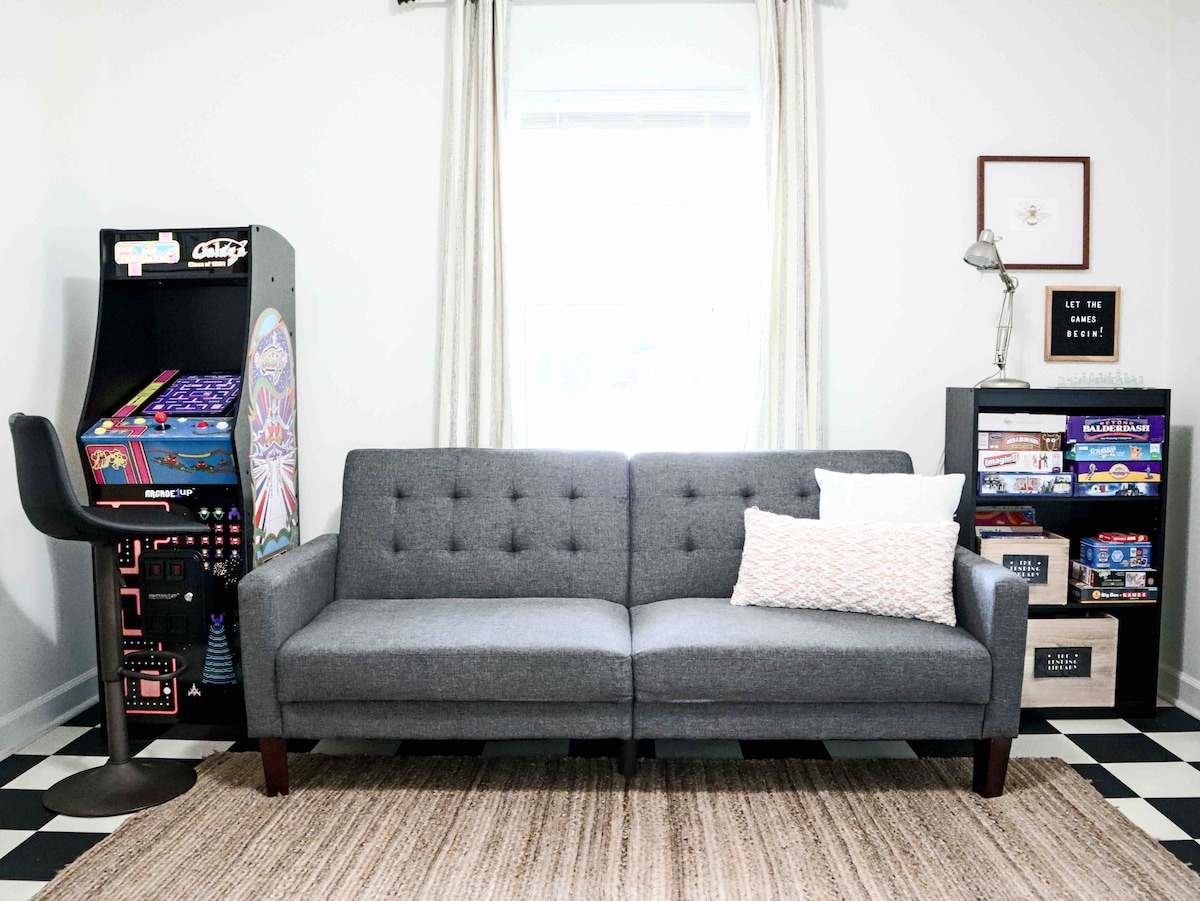 A modern living area is presented with a gray tufted sofa positioned against a light wall, complemented by a textured area rug. An arcade game machine stands nearby, alongside a bookshelf filled with games and books, while a sleek lamp adds a practical touch.