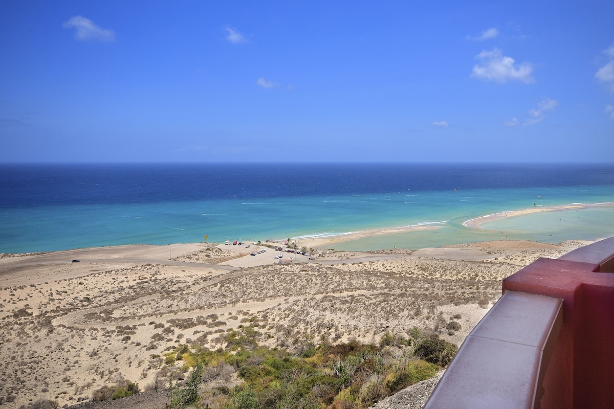 A panoramic view showcases the expansive coastline and the vibrant colors of the ocean. Soft sandy beaches blend into the azure water, while gentle waves create a serene atmosphere. The landscape is dotted with patches of greenery, providing a natural contrast to the beach and sea.