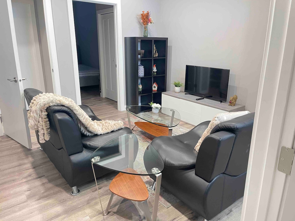 A cozy living area features two black leather armchairs facing a glass coffee table. A simple television stand holds a TV, while a bookshelf displays decorative items. Light-colored walls and hardwood flooring create a bright and inviting space.