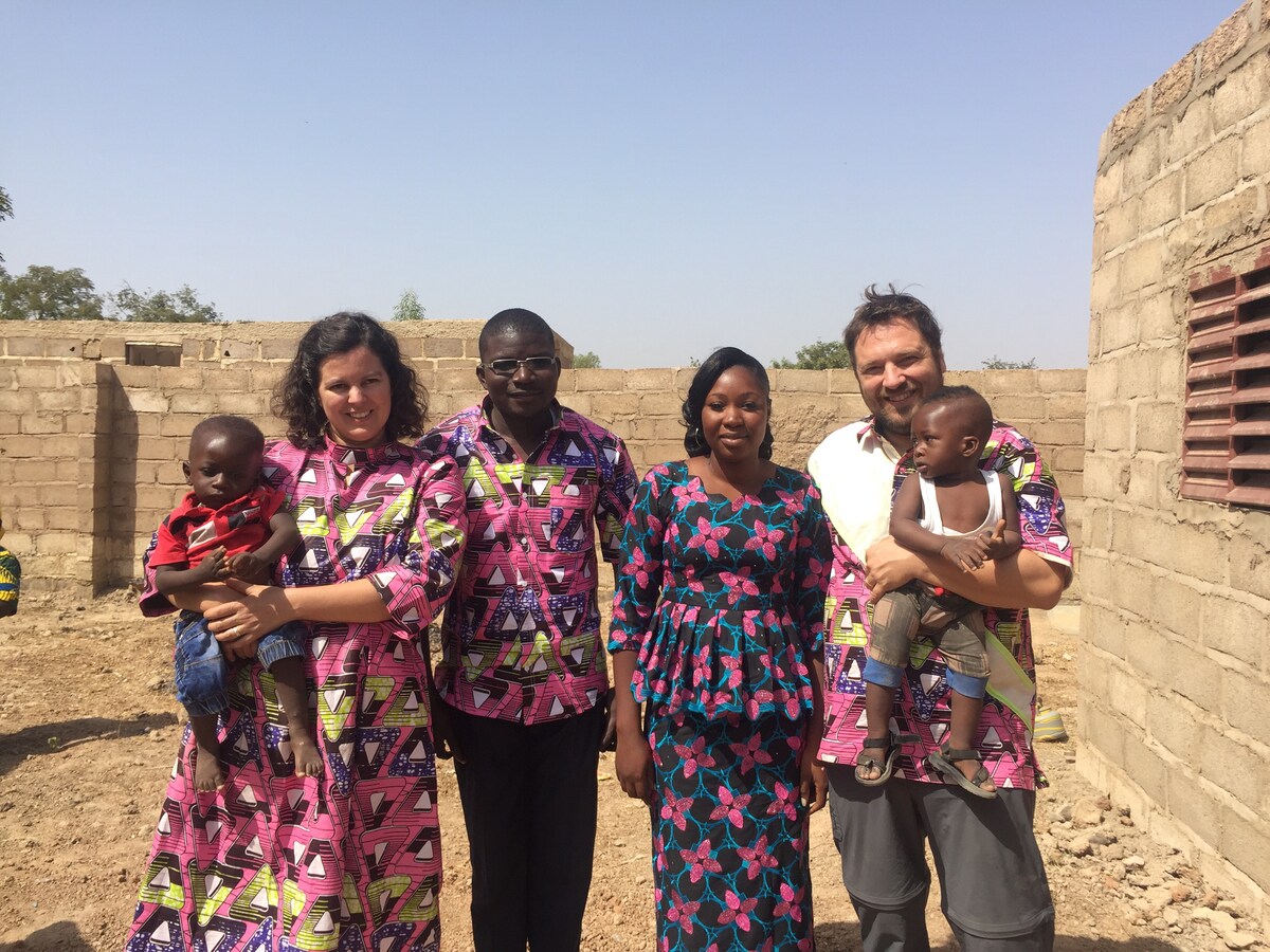 A group of five individuals stands outdoors against a backdrop of a stone structure. They wear matching colorful clothing, with two children being held. The scene conveys a sense of community and warmth in a rural setting.