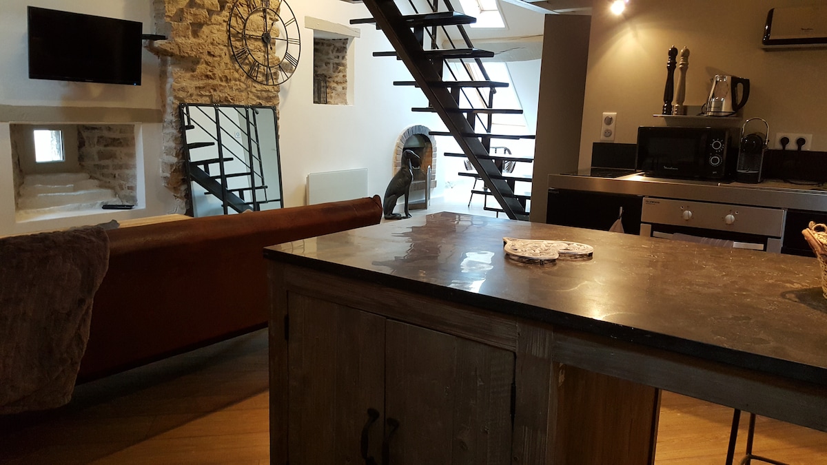 An open living space showcases a stylish kitchen island with a dark countertop. A modern spiral staircase leads to the mezzanine level, while exposed stone walls provide texture. A comfortable seating area is visible, complemented by a wall-mounted television and artistic decor.