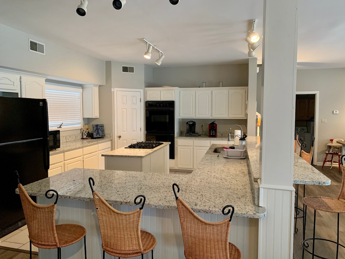 A spacious kitchen is equipped with light-colored cabinetry and granite countertops, featuring a central island with bar seating. Appliances include a black refrigerator and stove. Natural light enters through a window, and additional seating is available in the adjacent area.