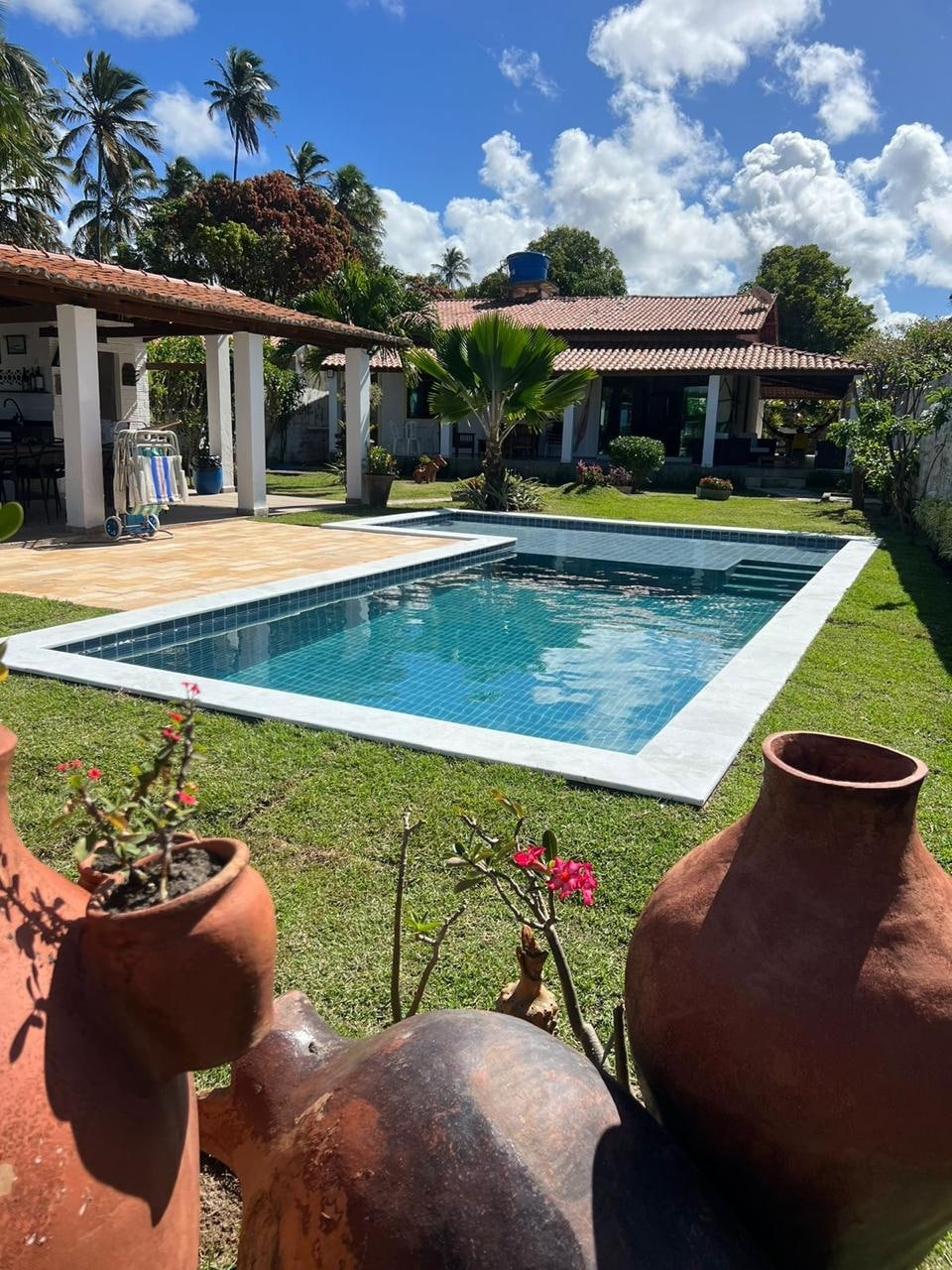 An inviting pool area is showcased, surrounded by lush greenery and colorful decorative pots. Smooth, clear water reflects sunlight, while a sunlit deck offers space for relaxation. The structure features a tiled roof, and a tropical garden enhances the serene atmosphere of the outdoor space.