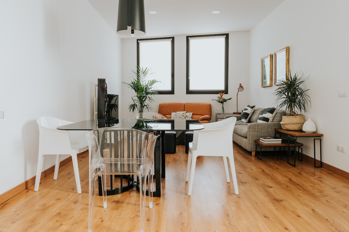 A spacious living area features hardwood flooring and large windows allowing ample natural light. A glass dining table is surrounded by modern chairs. A comfortable sofa and an accent chair are positioned near green plants, creating an inviting space for relaxation.