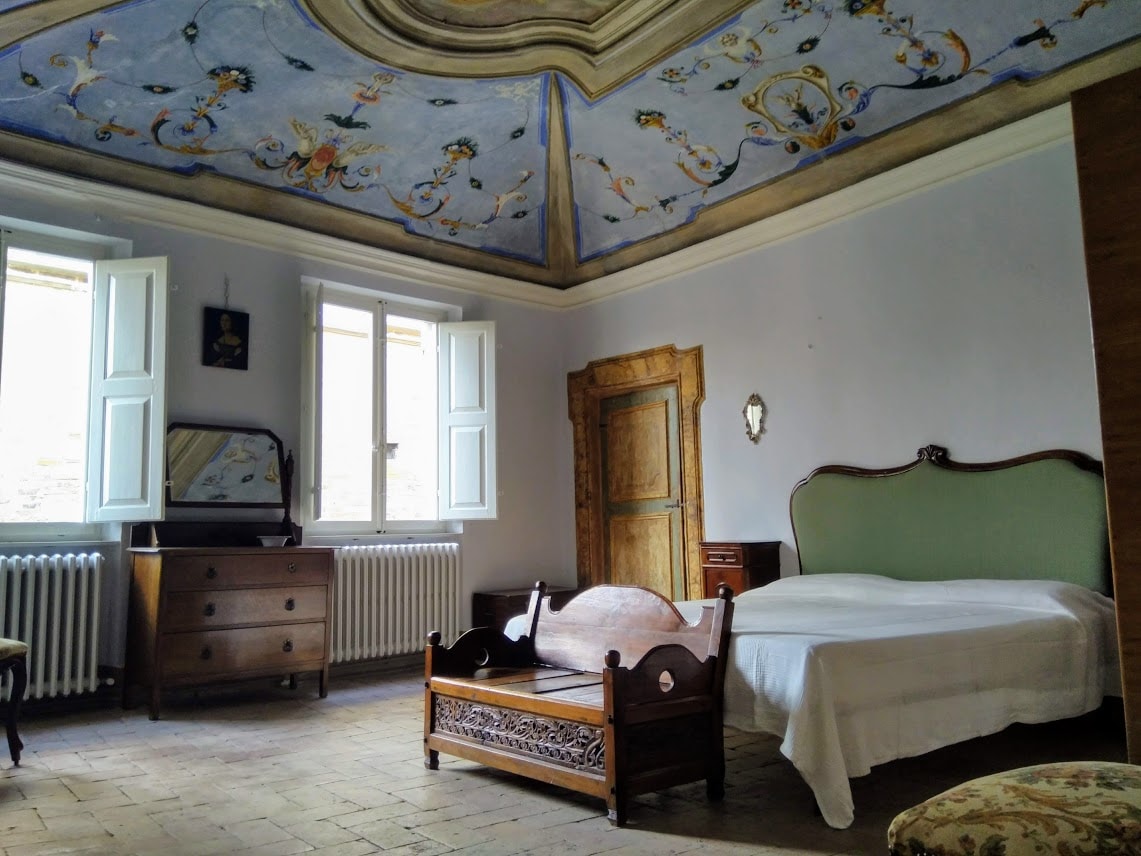 A spacious bedroom showcases a beautiful ceiling adorned with frescoes. Natural light floods in through two open windows, while a large bed is complemented by antique furnishings, including a wooden dresser and a decorative couch. The room features a blend of classic style and modern comfort.