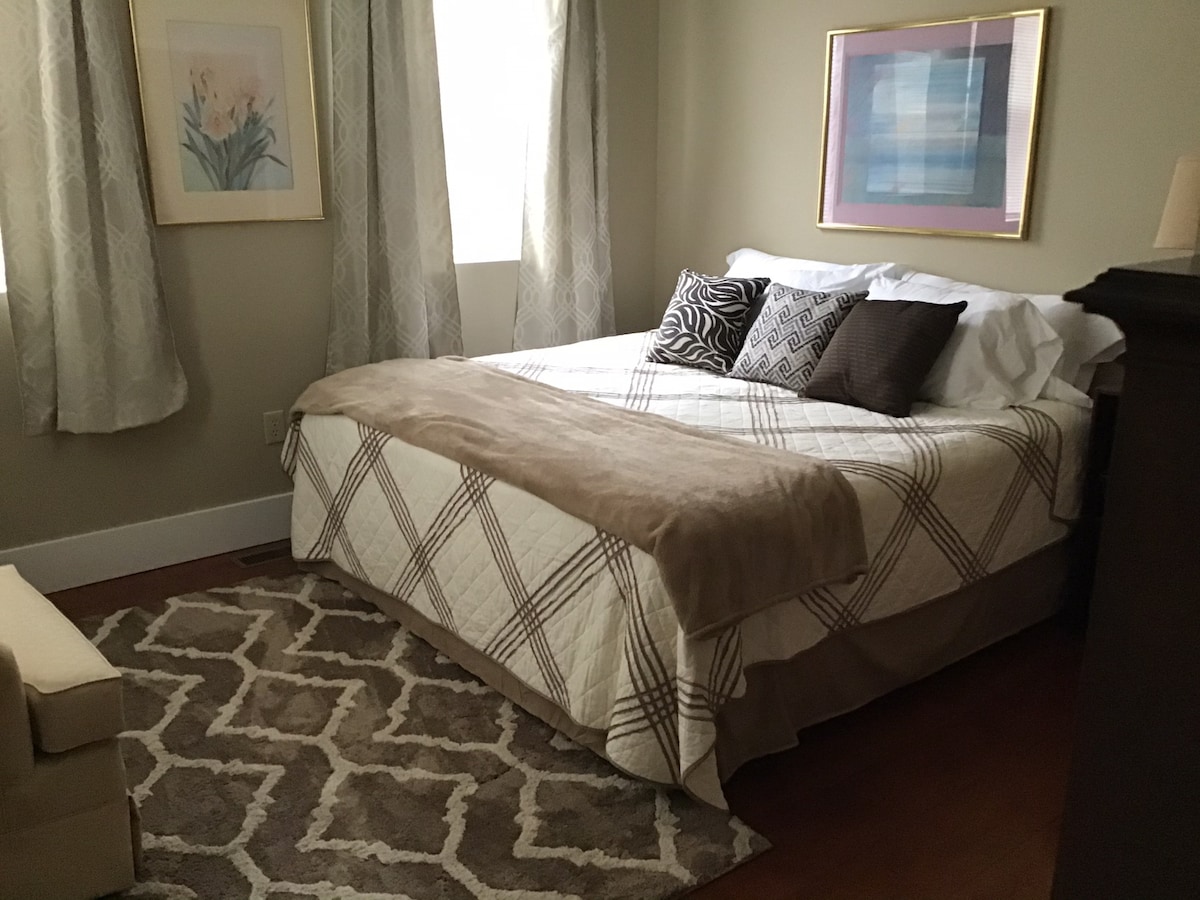 A cozy bedroom is presented with a spacious bed dressed in soft linens and decorative pillows. Natural light enters through partially draped windows, illuminating the inviting room. A plush rug and comfortable seating area complement the overall design.
