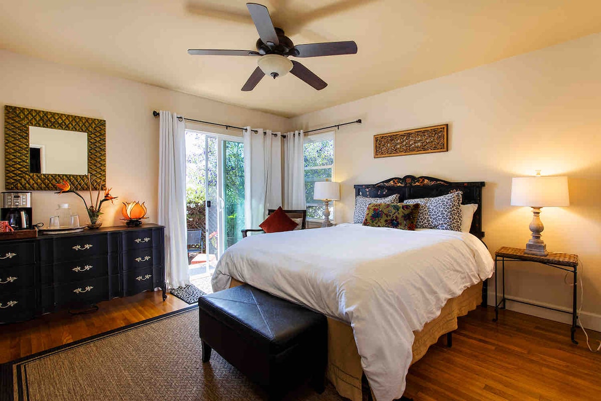 A cozy bedroom features a queen-sized bed with a light duvet and decorative pillows. A ceiling fan is located above, and light streams in through sliding doors that lead to an outdoor space. A dresser and nightstands complement the hardwood flooring.