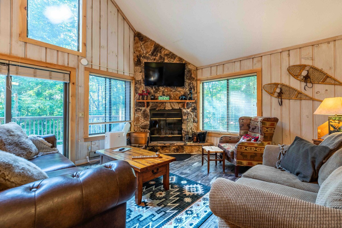 A cozy living area features a stone fireplace as a focal point, complemented by a mounted television above. Large windows provide a view of the surrounding greenery. Comfortable seating arrangements include a brown leather sofa and patterned armchairs, enhancing the cabin's relaxing atmosphere.