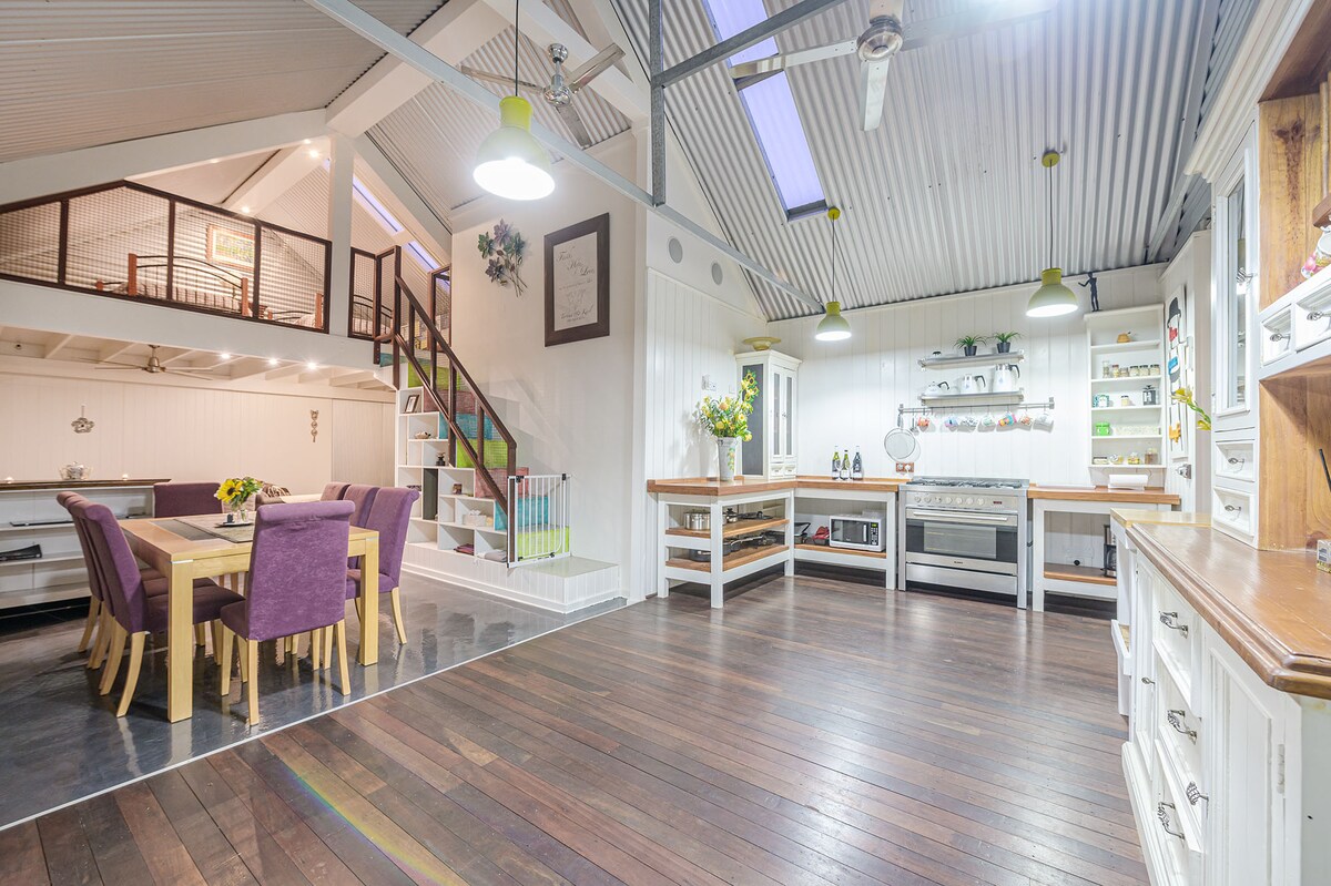 An open living area features polished wooden floors and high ceilings with skylights, allowing natural light to fill the space. A fully equipped kitchen is displayed with various cooking utensils, while a dining table is surrounded by upholstered chairs. A staircase leads to a loft area, enhancing the home's spaciousness.
