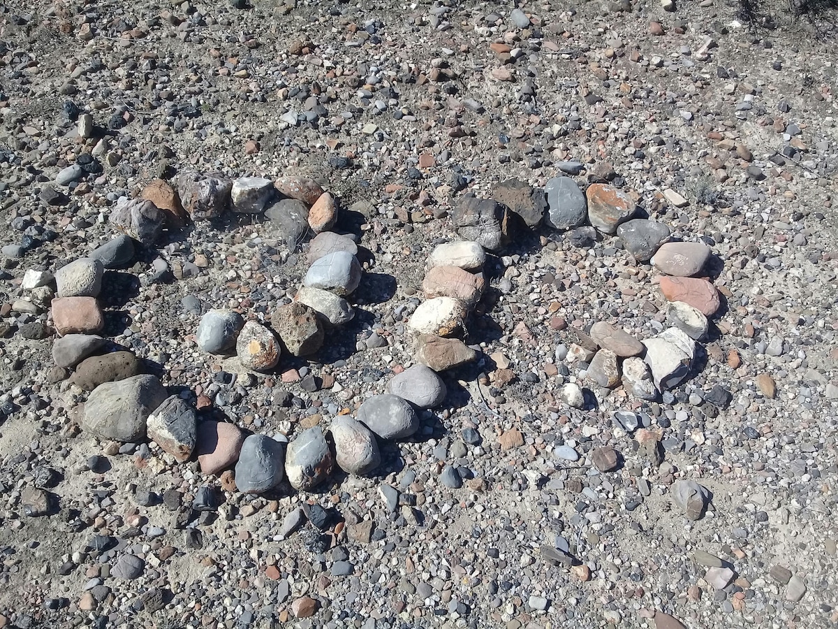 A circular arrangement of various sized stones is visible on a gravelly desert ground. The stones form two distinct shapes, creating a natural design that blends with the arid surroundings.