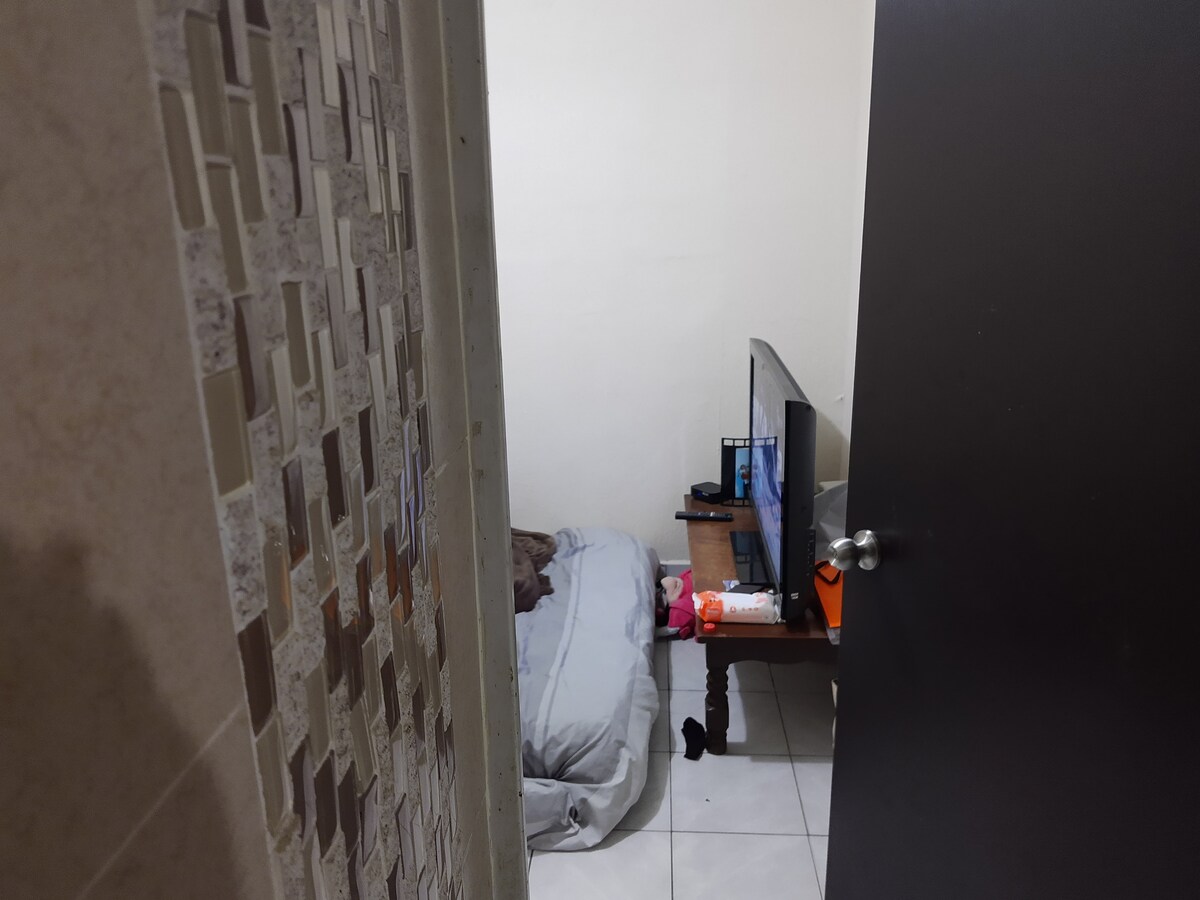 A small cozy room is visible through an ajar door. A spacious bed is placed against one wall, while a wooden table stands nearby, supporting a television and personal items. Neutral-colored walls and tiled flooring create a simple, functional environment.