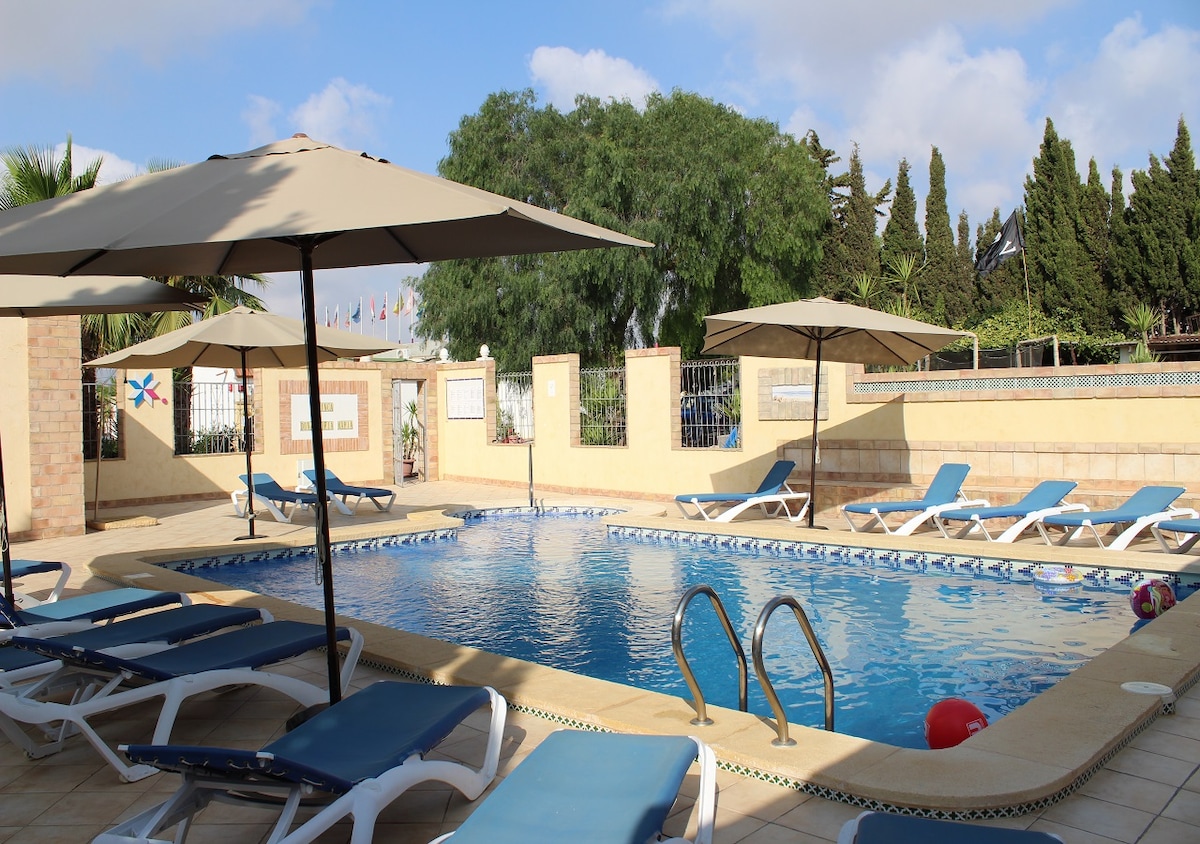 The communal pool area features a clear blue pool, surrounded by multiple sun loungers and large umbrellas for shade. Lush greenery lines the background, creating a relaxed environment for guests. The pool steps provide easy access for a refreshing swim.