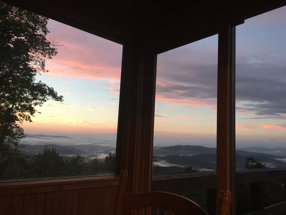 A serene view of the mountains is captured through large windows, displaying layers of hills fading into the distance. Soft pink and orange hues fill the sky during sunrise, while clouds hover gently over the valleys. The scene evokes a sense of tranquility.