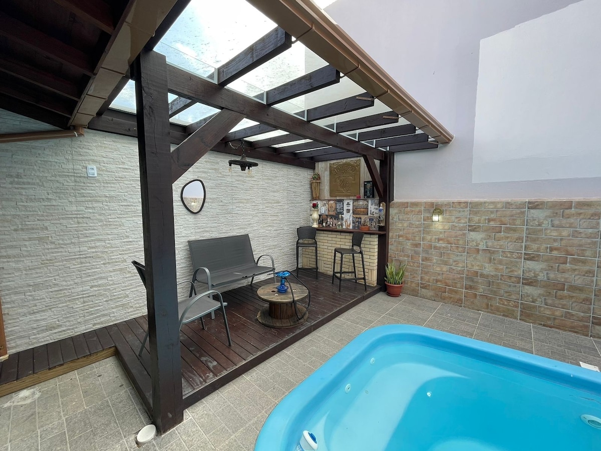 A covered outdoor area features a small pool and inviting seating arrangements. A table is positioned between two chairs, with a bar-like structure in the background. The space is complemented by wooden beams and a textured stone wall, providing an open yet cozy atmosphere.