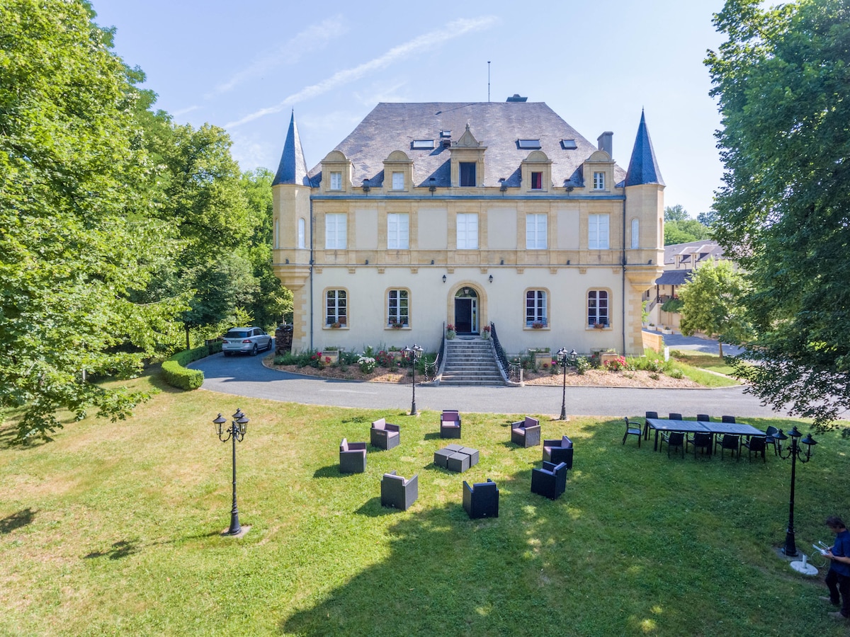 The exterior of a historic château is depicted, featuring a grand façade with tall windows and pointed towers. A landscaped garden surrounds the building, showcasing neatly arranged seating areas with dark chairs on the lawn, complemented by decorative lampposts creating a welcoming outdoor space.