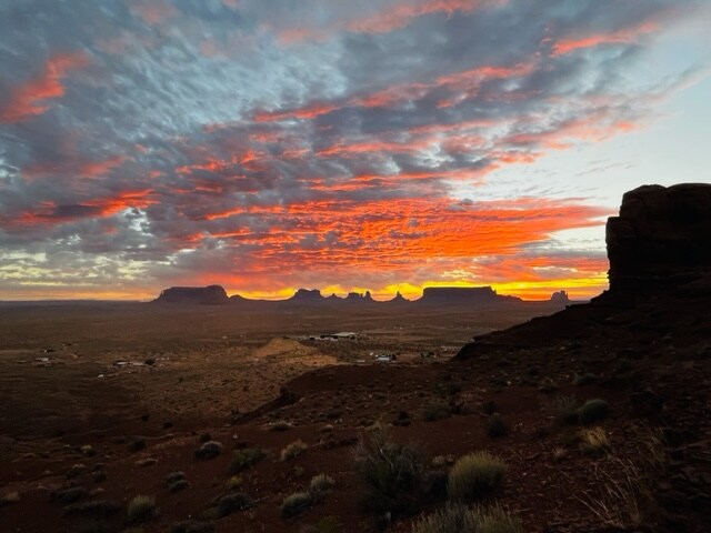 A vibrant sunset casts rich hues of orange and red across a vast desert landscape. Silhouetted rock formations and mesas create a striking horizon, while rolling terrain dotted with shrubs adds texture to the scene. The sky is filled with dramatic clouds, enhancing the natural beauty.