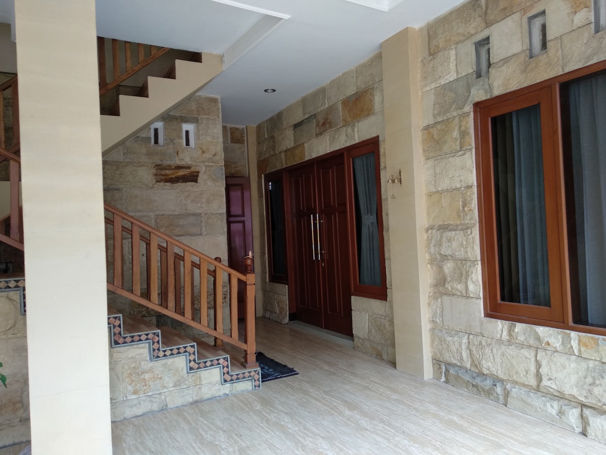 The entrance area features natural stone walls and wide windows covered with sheer curtains. A wooden staircase leads to the upper floors, while a double-door entrance displays elegant detailing. The flooring is finished in light-colored tiles, enhancing the spacious feel.