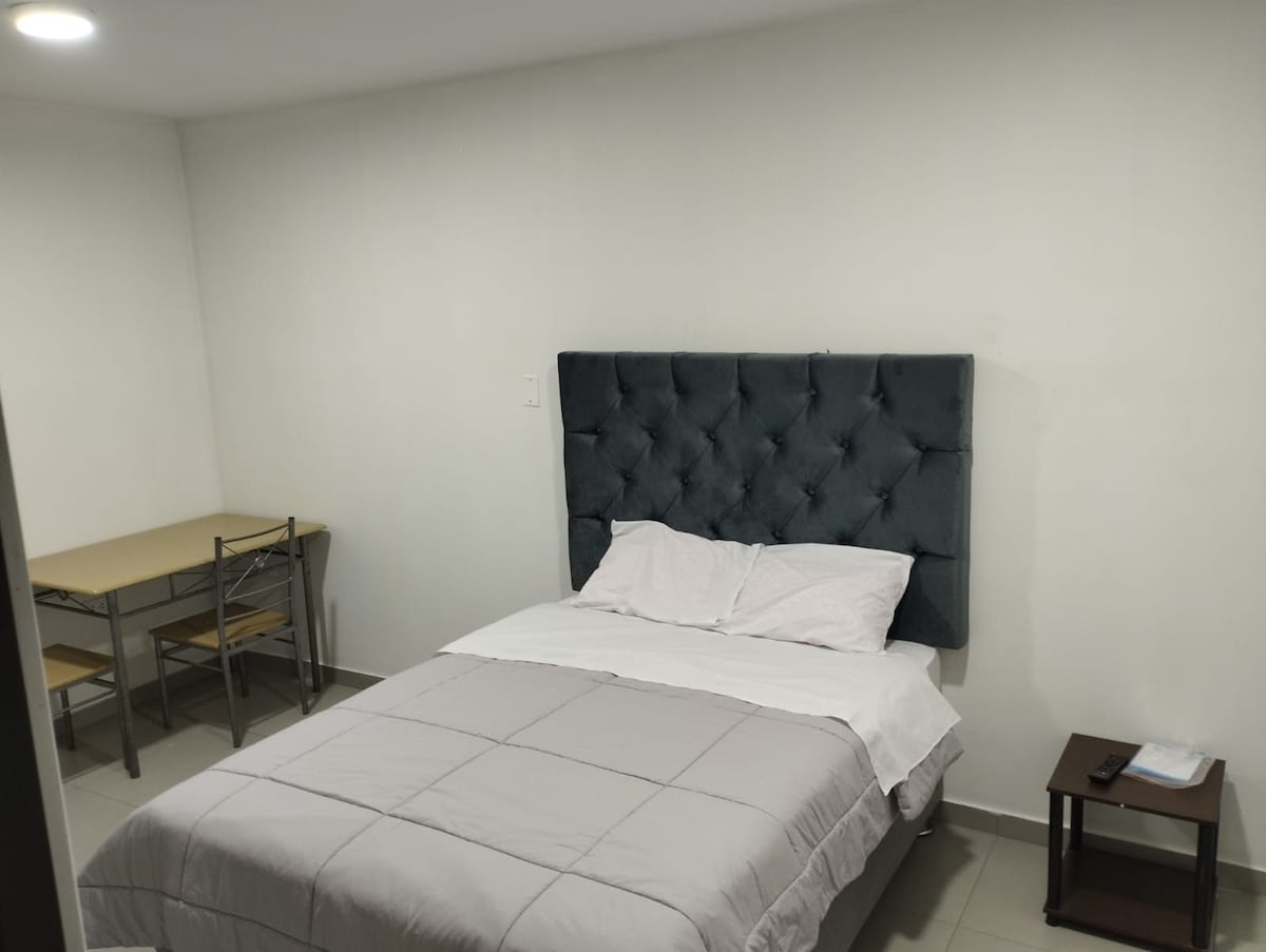 A simple sleeping area features a spacious bed with a tufted headboard and light bedding. A small wooden table with two chairs is positioned against the wall, alongside a dark side table providing additional surface space. The room is lit with soft overhead lighting.