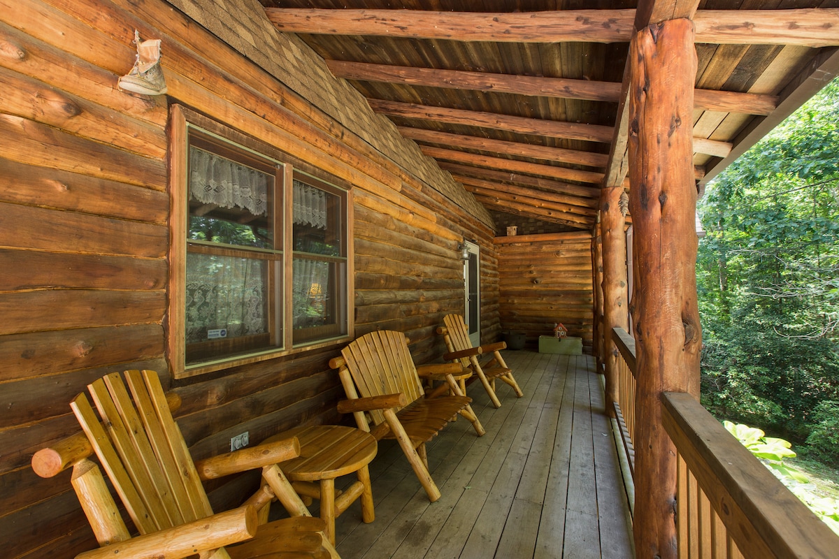 A covered porch features several rustic wooden rocking chairs arranged along the length, offering a space for relaxation. The log cabin's natural wood exterior is visible, surrounded by greenery, providing a connection to the outdoors.
