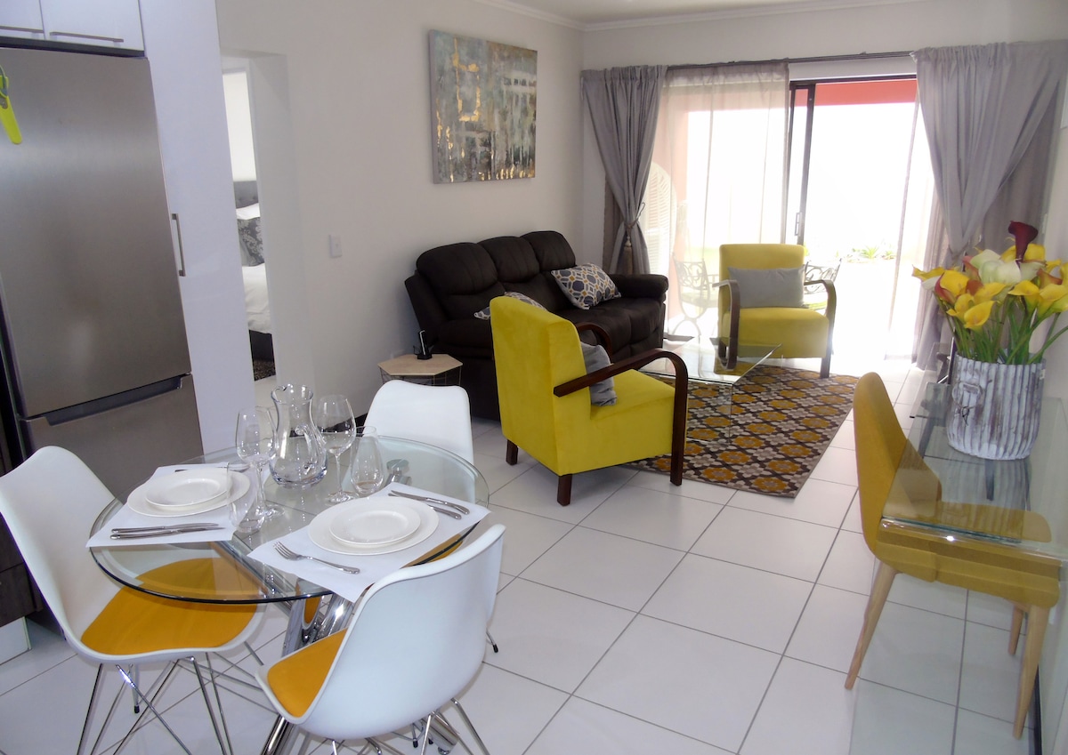The living area features a leather couch and two accent chairs, complemented by a glass coffee table. A dining table set for four is positioned nearby. Natural light floods in from sliding glass doors that lead to an outdoor space, enhancing the open feel of the room.