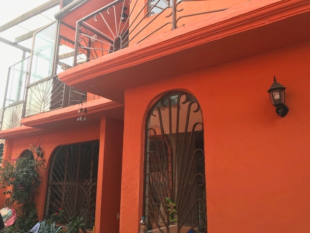 The exterior of a two-story house is visible, featuring an orange facade with decorative wrought-iron railings and window grilles. Several plants are situated at the base of the entrance, contributing to a welcoming appearance. A lantern is mounted near the entrance, adding to the charm.