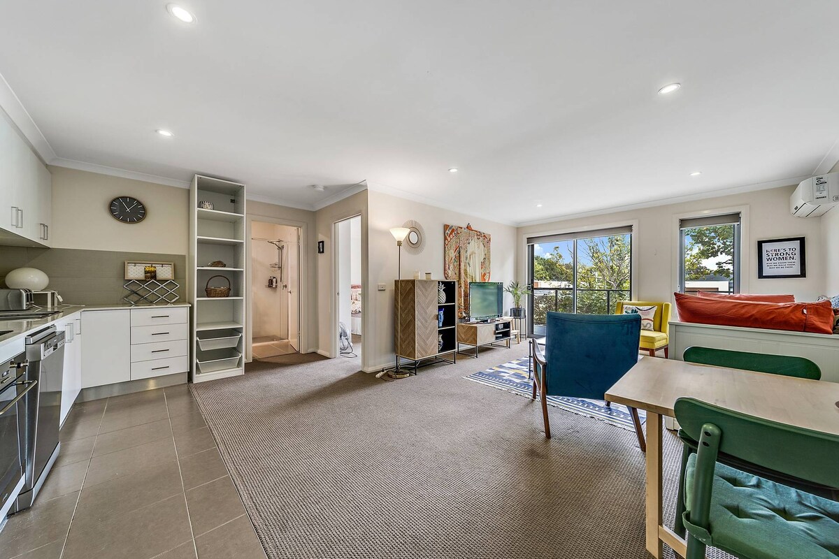 A well-lit open living area features stylish furnishings, including a multi-colored seating arrangement and a dining table. A flat-screen television is positioned for easy viewing. Large windows offer greenery views and allow natural light to illuminate the space.