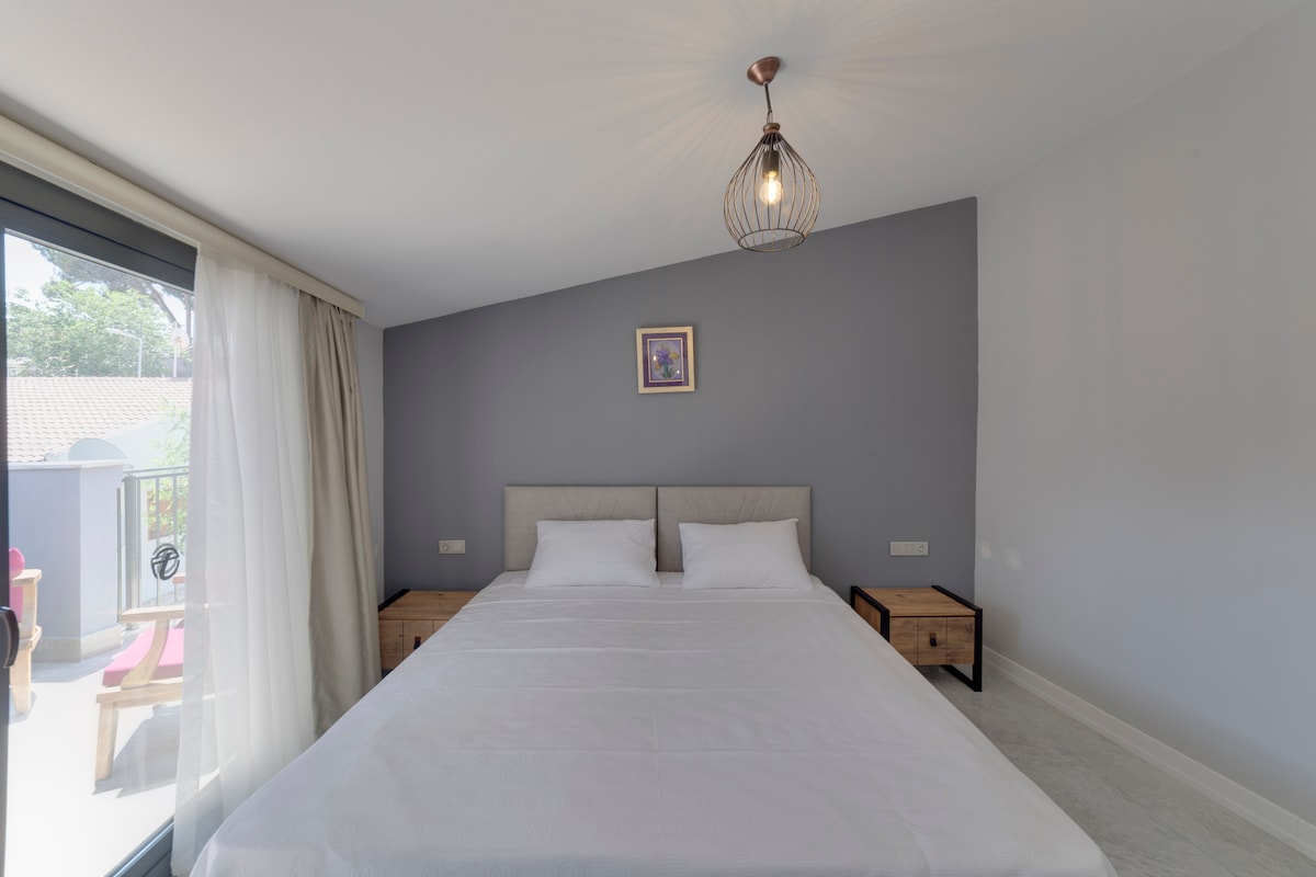 A spacious bedroom features a comfortable bed with light bedding and a neutral-colored headboard. Two wooden bedside tables are positioned on either side. Natural light enters through a large glass door, leading to a private balcony, while a modern light fixture hangs from the ceiling.