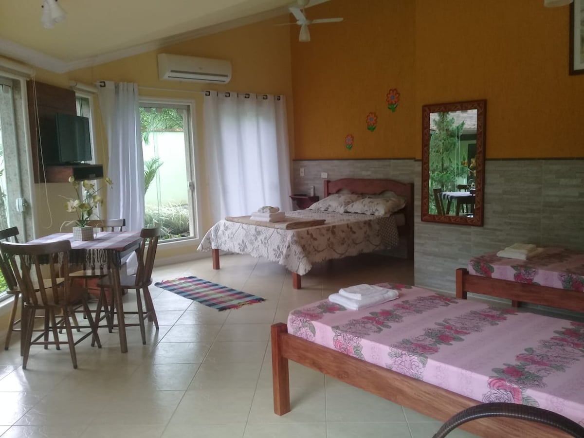 The spacious suite features a king-sized bed and two single beds arranged in a bright, open area. A wooden dining table with four chairs sits nearby. Natural light enters through large windows, revealing greenery outside, while a TV is mounted on a wall opposite the beds.