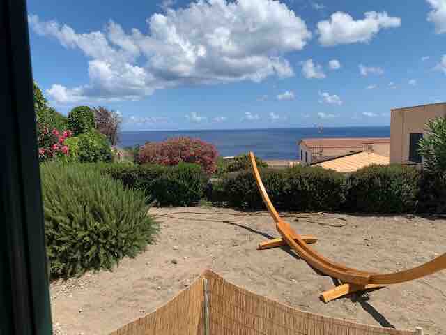 A serene outdoor space is visible, featuring a wooden hammock frame set on sandy ground, surrounded by low shrubs and flowers. The expansive view captures the shimmering sea under a partly cloudy sky, with neighboring buildings subtly in the background.