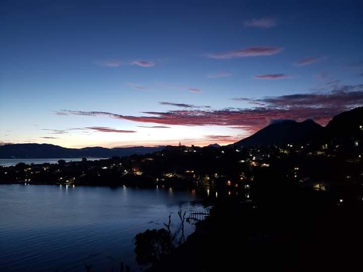 A serene evening view showcases the tranquil waters of Lake Atitlán, with silhouettes of nearby mountains under a twilight sky. Soft hues of pink and blue reflect in the calm surface, while lights from the town shimmer along the shore.