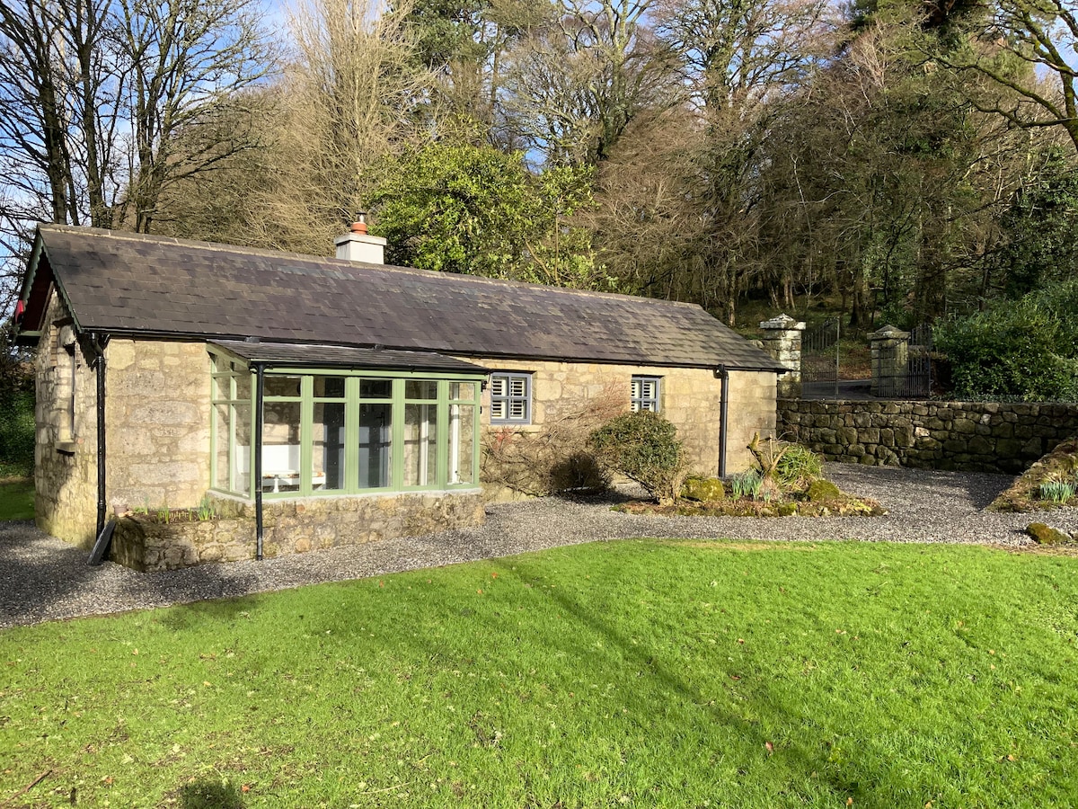 The cottage is framed by a lush green lawn, featuring a stone facade and a welcoming sunroom with large windows. Surrounding trees provide a natural backdrop, while a gravel pathway leads to the entrance. A stone wall delineates the property, enhancing its secluded feel.