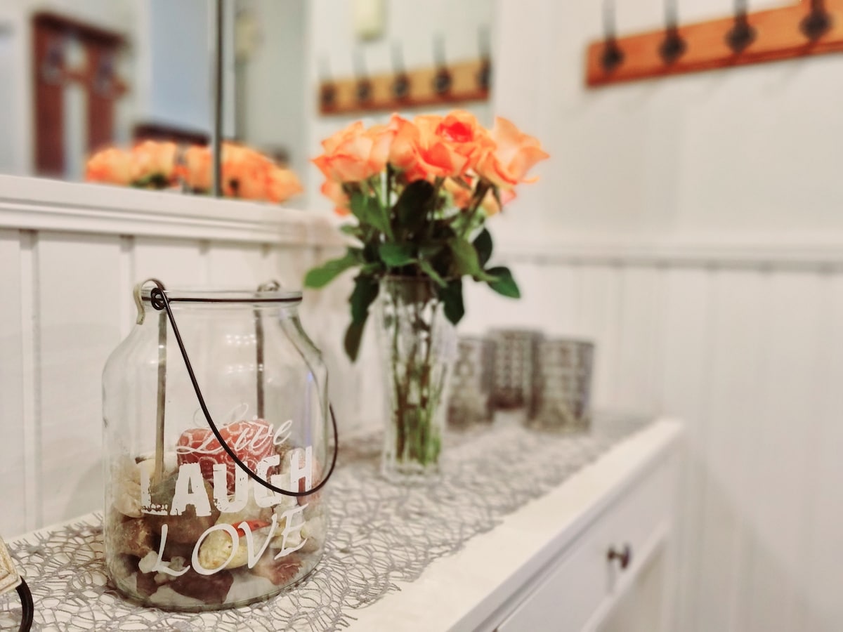 A welcoming entryway console table is adorned with a floral arrangement of orange roses. A glass jar containing decorative stones sits in front, alongside several small candle holders. The space features light-colored walls and a mirror that enhances the bright atmosphere.