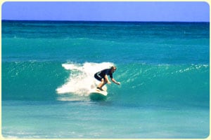An individual is seen riding a wave on a surfboard, with the ocean stretching in varying shades of blue. The surfer is positioned toward the crest of the wave, while foam can be observed breaking around them, contributing to the dynamic scene.