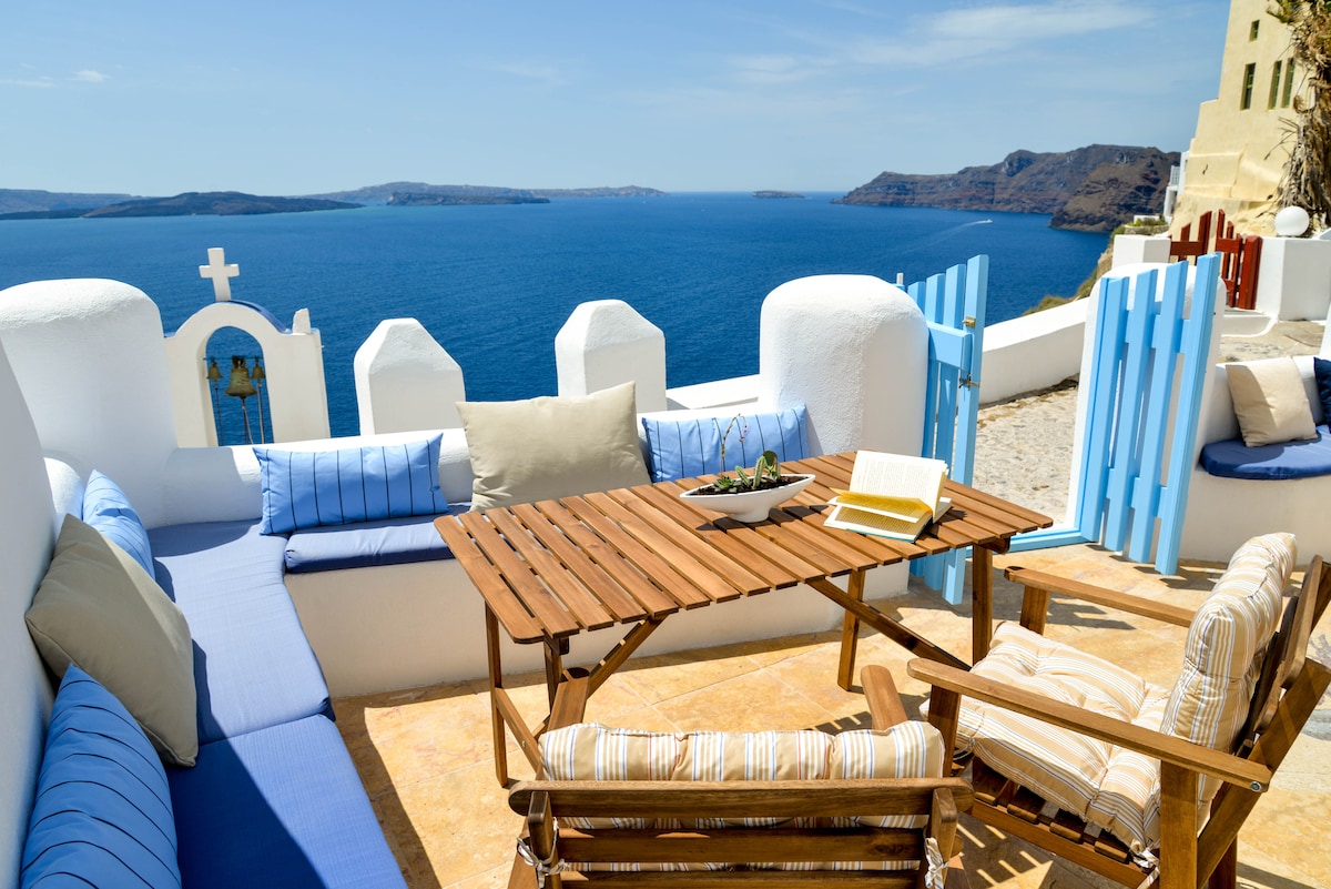 A private veranda is displayed with comfortable seating arrangements, including a wooden table surrounded by cushioned benches. The view overlooks the calm Aegean Sea and distant cliffs, while the light blue decorative gates add a charming touch to the tranquil atmosphere.