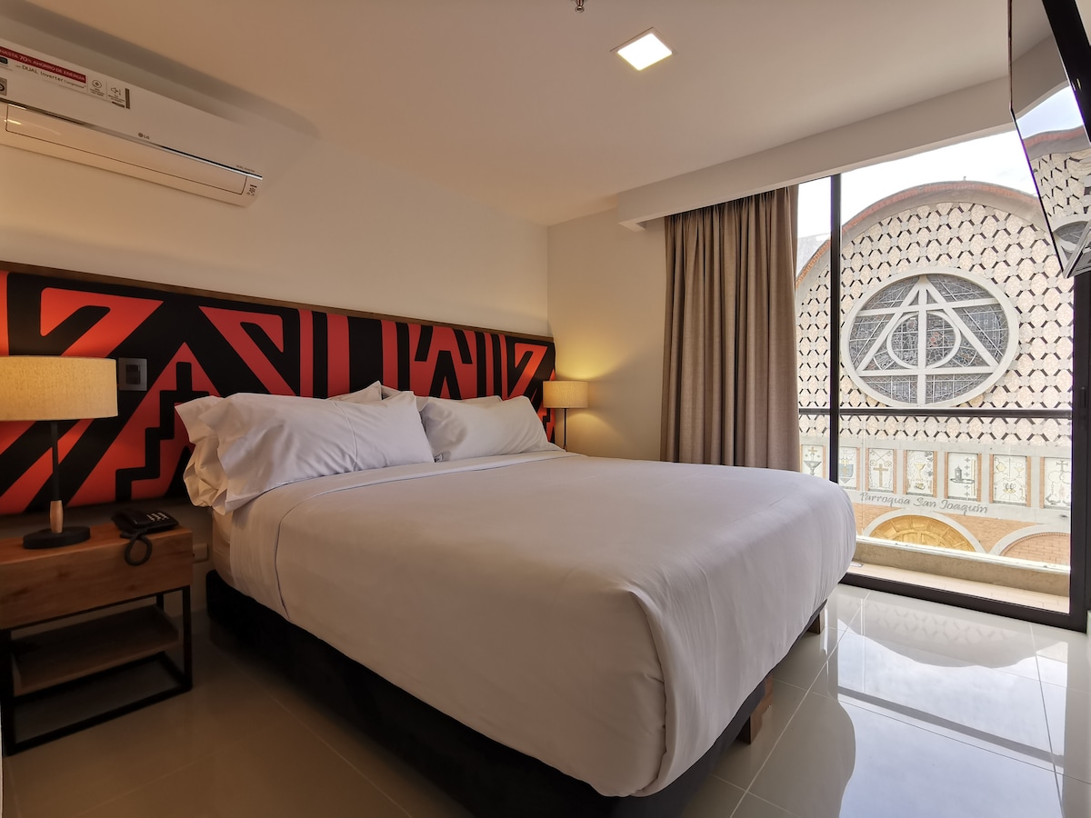 A spacious bedroom features a large king-sized bed with a modern headboard. Natural light streams through the large window, revealing an architectural view of the building outside. A soft lamp and bedside table complement the minimalist decor.