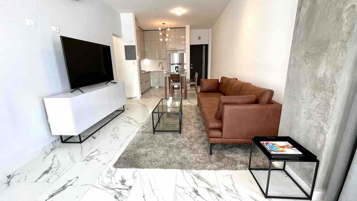 A modern living area is furnished with a brown leather couch, a coffee table on a soft rug, and a TV mounted on the wall. Light-colored marble flooring contrasts with the sleek cabinetry and kitchen area in the background, providing an open and airy feel.